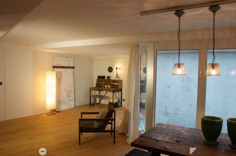 A cozy, well-lit room with wooden flooring, a hanging pendant light, a wooden table with vases, a comfortable chair, a desk area with a chair and lamp, and a large window with sheer curtains.