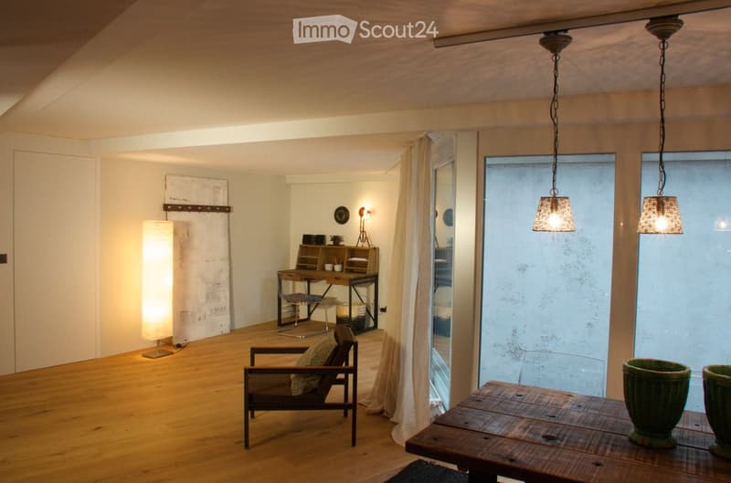 A cozy, well-lit room with wooden flooring, a hanging pendant light, a wooden table with vases, a comfortable chair, a desk area with a chair and lamp, and a large window with sheer curtains.