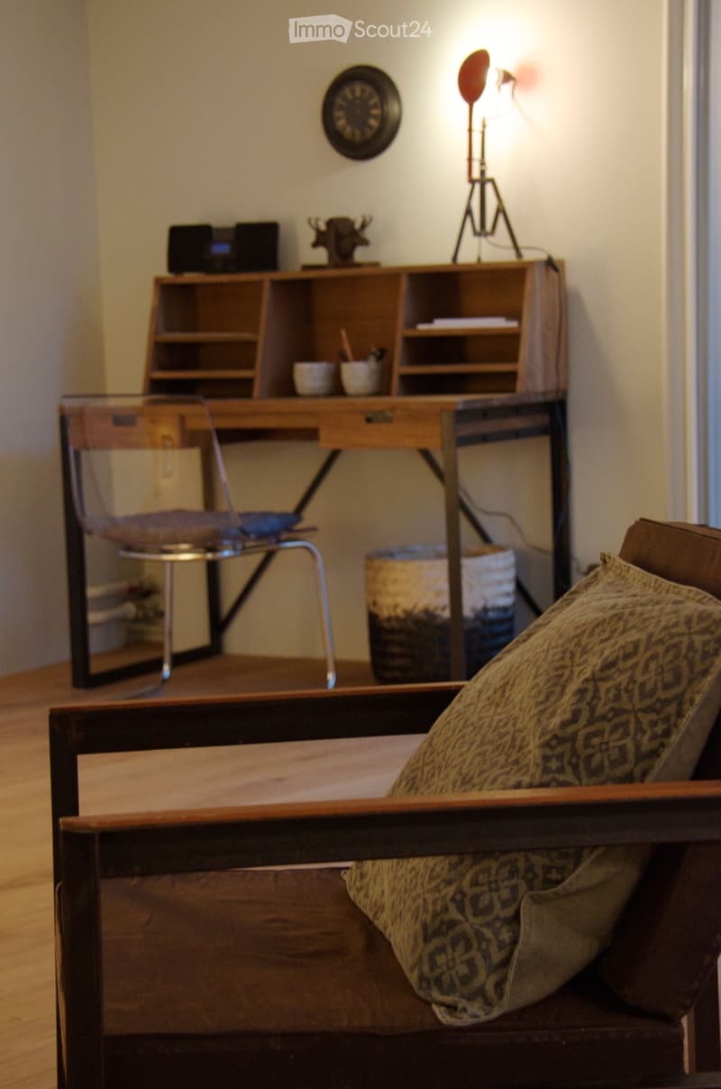 A cozy living space with a wooden sofa, patterned cushions, a glass-top chair, a wooden desk with a shelf, and a wall-mounted clock.
