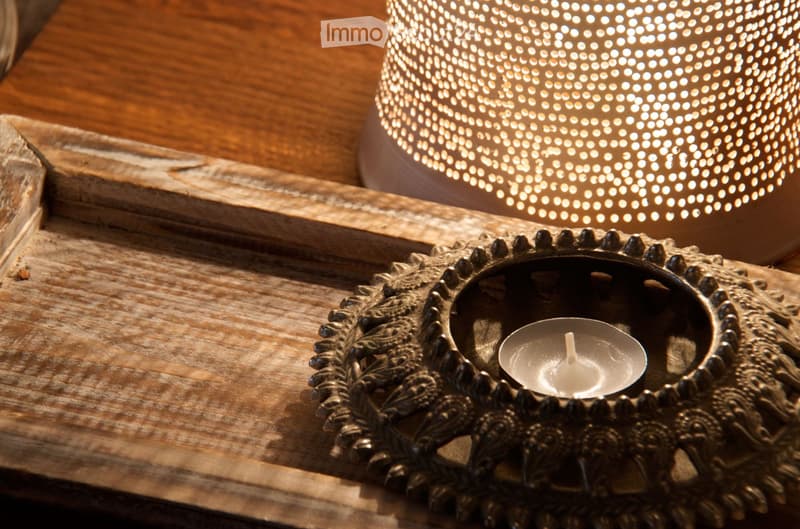 A wooden table with a decorative candle holder and a lit candle, alongside a round, perforated candle holder with small lights.