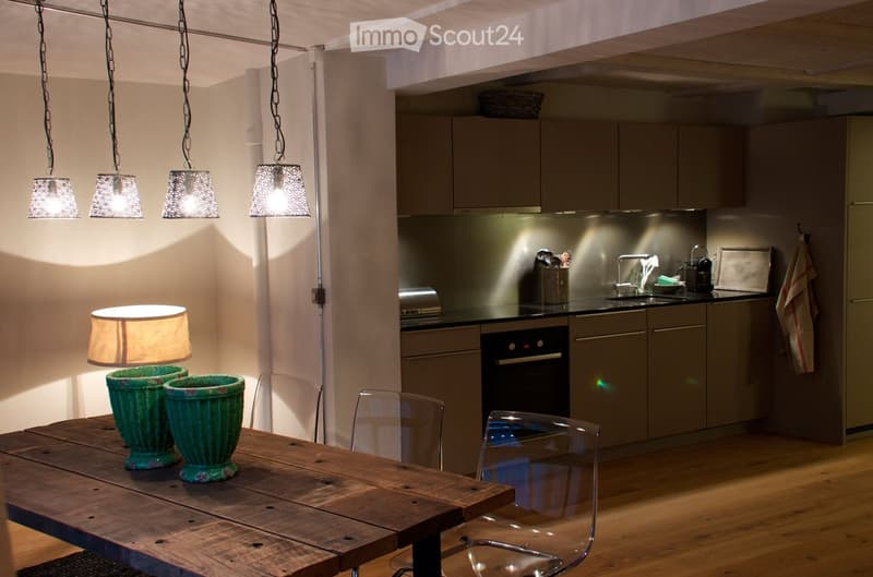 Open-plan kitchen and dining area with wooden flooring, modern cabinets, stainless steel appliances, a wooden dining table with two green vases, and hanging pendant lights.