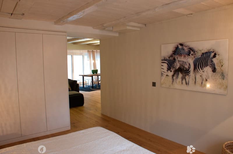Bright bedroom with wooden flooring, a large bed, a modern wardrobe, and a large painting of zebras on the wall. An open doorway leads to a living area with a sofa, table, and chairs.