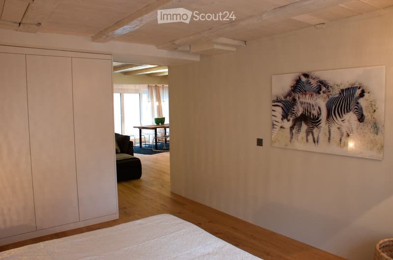 Bright bedroom with wooden flooring, a large bed, a modern wardrobe, and a large painting of zebras on the wall. An open doorway leads to a living area with a sofa, table, and chairs.