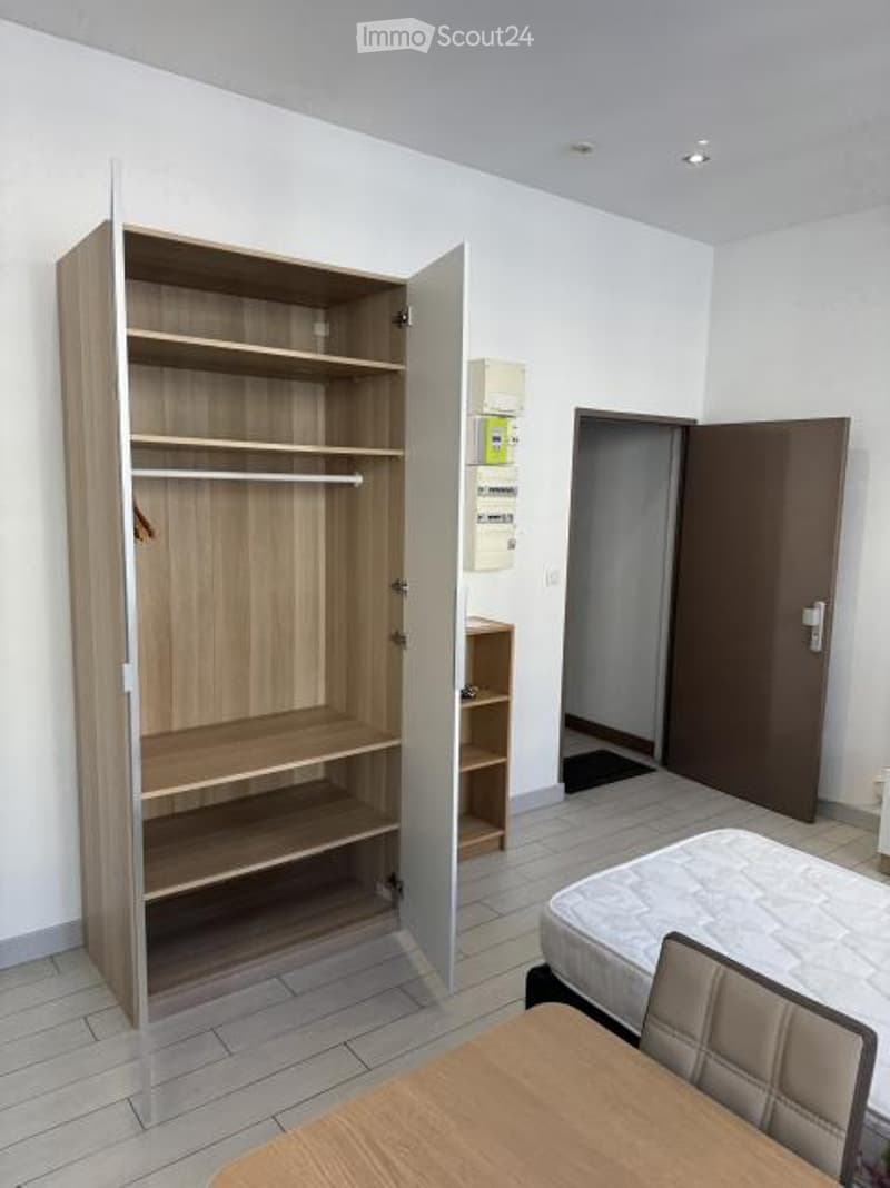 A furnished room with a wooden wardrobe, a bed, a small table, and a chair. The room has tiled flooring and white walls.