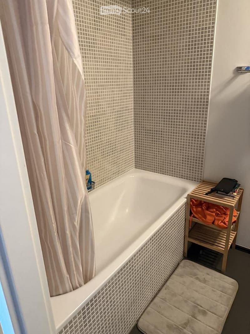 Bathroom with a white bathtub, beige tiled walls, a wooden shelf holding a bathrobe, and a beige bath mat on the floor.