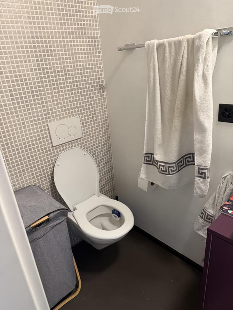 Modern bathroom with white tiled walls, a toilet, a towel hanging on a rack, and a small cabinet.
