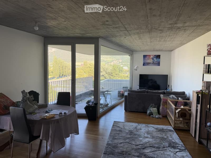 Open-plan living and dining area with large windows offering a scenic view, featuring a sofa, dining table with chairs, television, and a floor lamp.