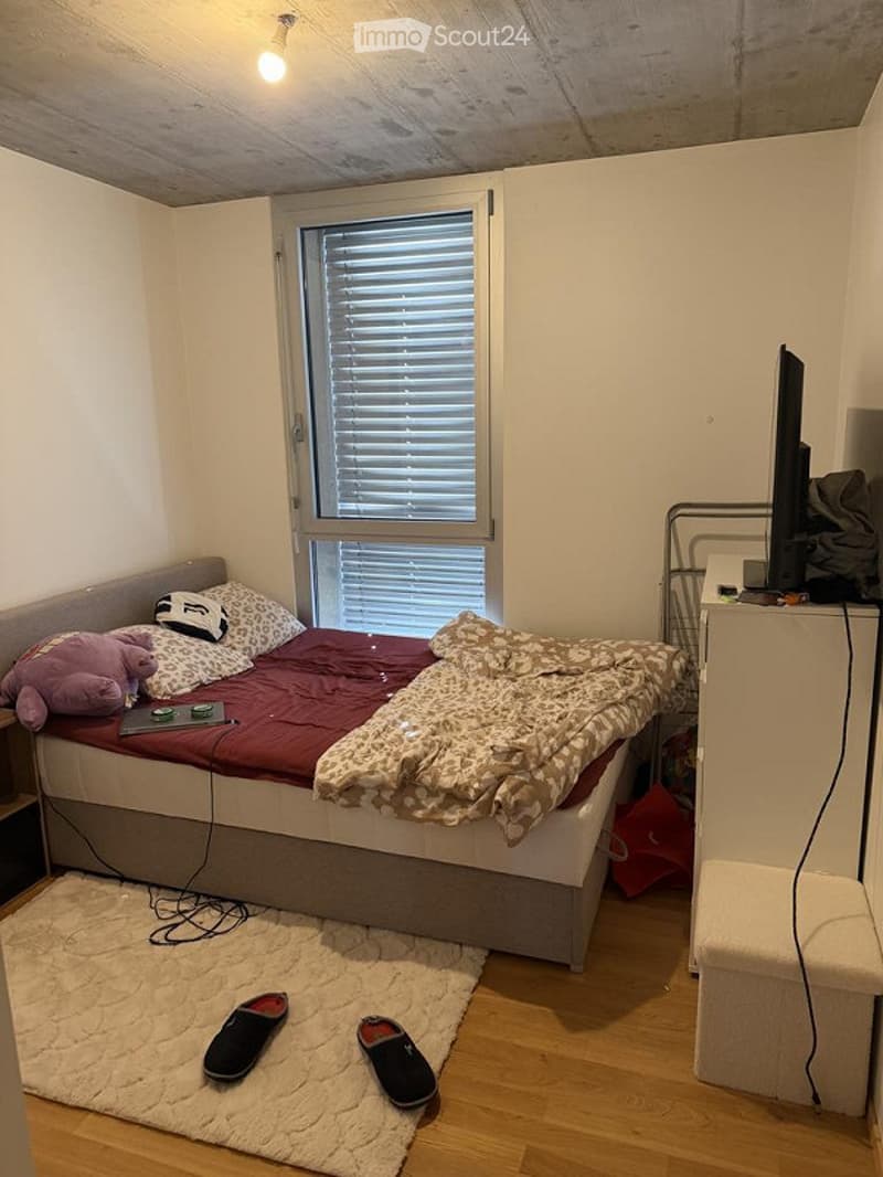 Cozy bedroom with a single bed, pillows, blanket, laptop on the bed, a small stool, a screen, and slippers on the floor.