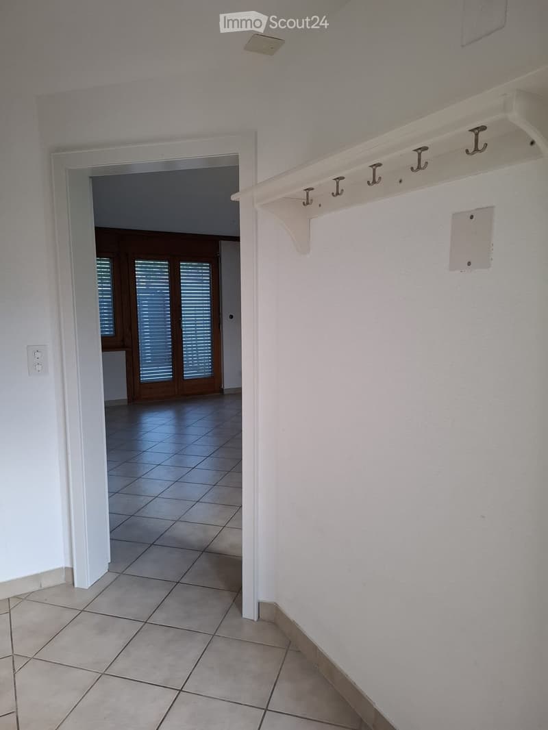 Empty hallway with tiled floor, white walls, and a coat rack with hooks on the right side. An open doorway leads to another room with wooden double doors and blinds.
