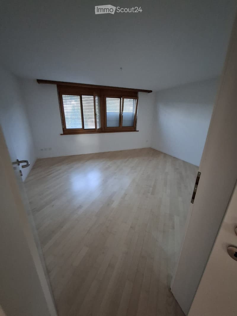 Empty room with wooden flooring and white walls, featuring a window with wooden frame and blinds, and a door with a silver handle.