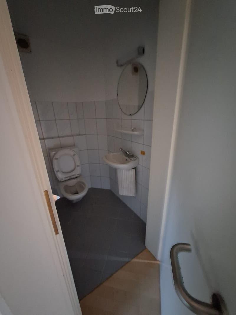 Small bathroom with a toilet, sink, and mirror. The walls are tiled, and the floor is a light brown material.