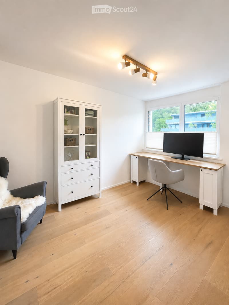 Bright, modern room with light wooden flooring, white walls, and large windows. Features a grey sofa with a white throw, a white cabinet with glass doors, a wooden desk with a screen, and a sleek chair.