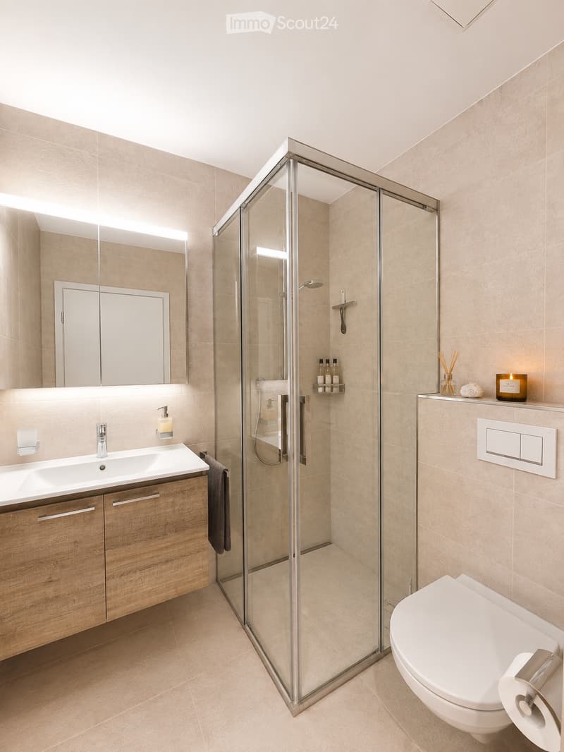 Modern bathroom with a glass-enclosed shower, wooden vanity with sink, mirror, and toilet. Features include a towel, candle, and decorative items on the shelf.