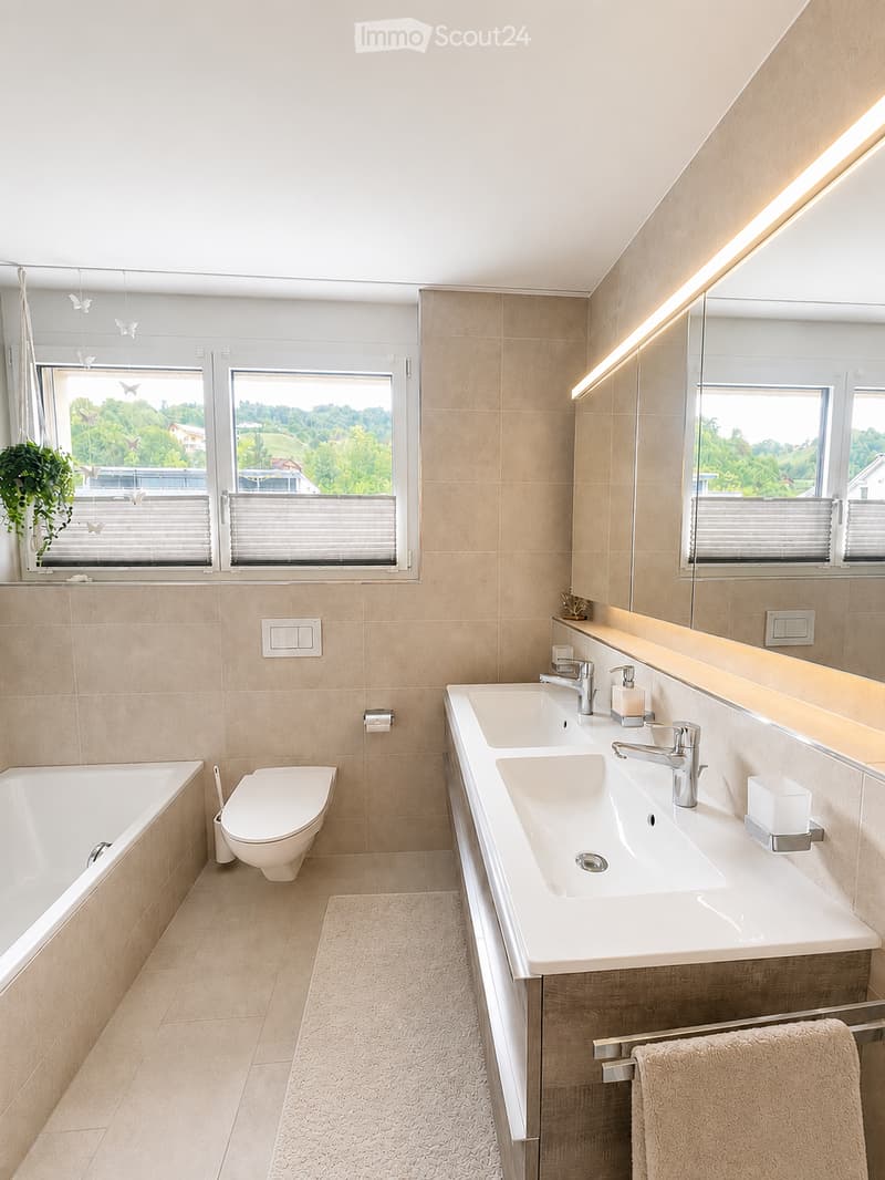 Modern bathroom with beige tiles, a large mirror, double sink, bathtub, toilet, and a window with blinds offering a scenic view.