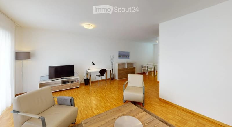 Bright living room with wooden flooring, white walls, and large windows. Features a beige sofa, coffee table, TV stand, floor lamp, and a small dining area with chairs and a table.