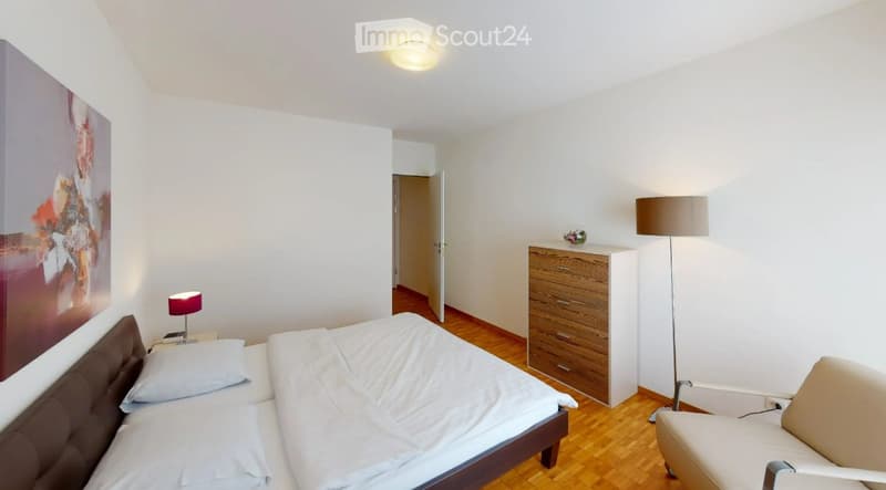 A modern, well-lit bedroom with a double bed, nightstand, lamp, chair, drawer unit, and a large picture on the wall.
