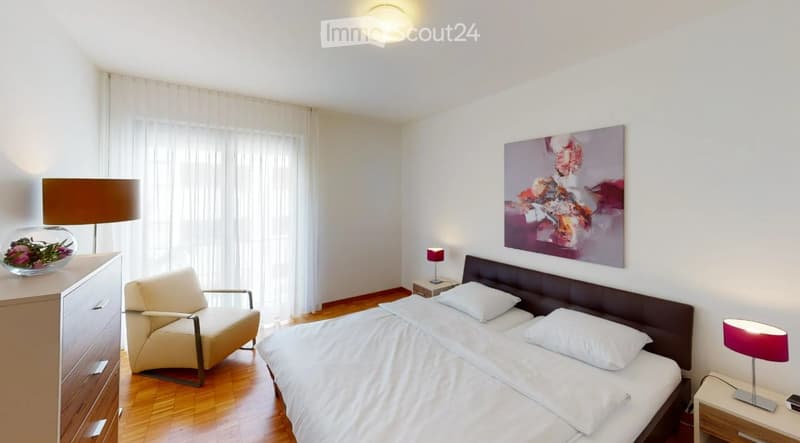 Modern bedroom with a large bed, nightstands, a wooden dresser, a chair, and a decorative vase on the dresser. The room features large windows with white curtains and a colorful abstract painting on the wall.