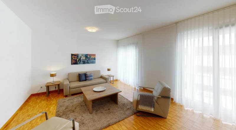Bright living room with wooden flooring, large windows with white curtains, a beige sofa with two throw pillows, a wooden coffee table with a decorative plate, an armchair with a throw blanket, side tables with lamps, and a picture frame on the wall.
