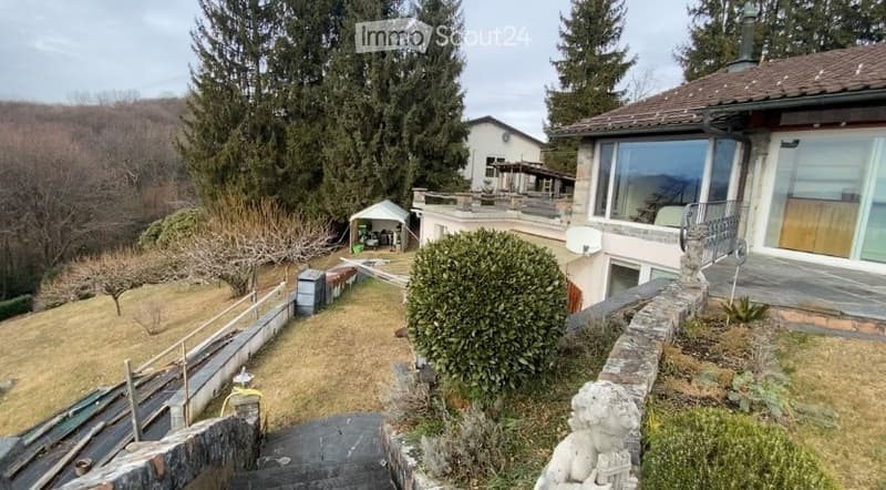 A well-maintained garden area with a stone pathway leading up to a house. The garden includes neatly trimmed hedges, a small gazebo, and a stone wall bordering the property. The house features large windows and a modern design, with a view of surrounding