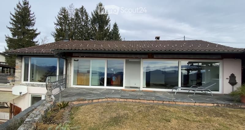 Single-story house with large glass sliding doors, stone patio, and outdoor lounge chairs.