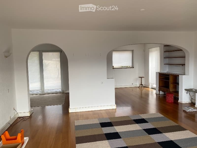 Bright, open-concept living space with wooden flooring, large windows, and arched doorways. Features a colorful geometric rug, a small wooden stool, a wooden cabinet, and a small table with a chair.