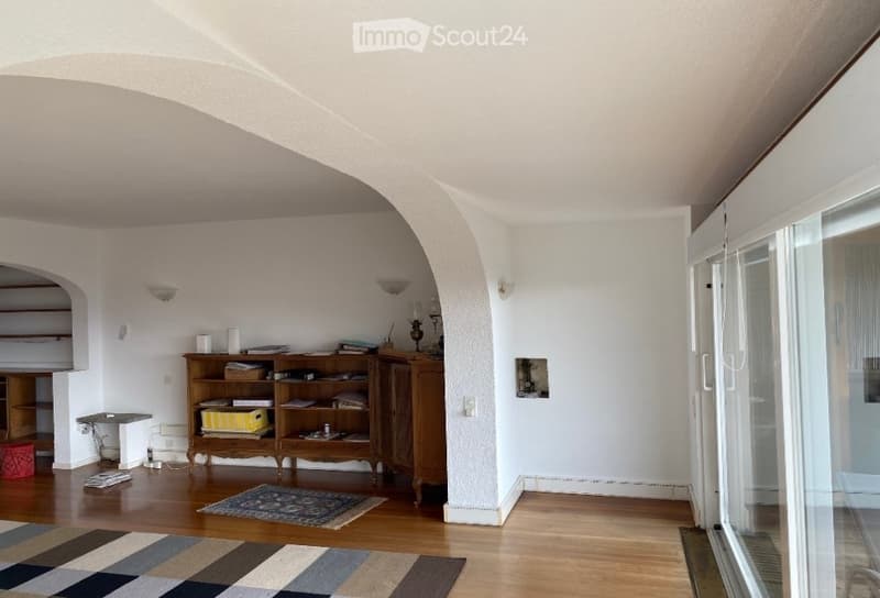 Bright, open living space with wooden flooring, white walls, arched doorways, and large windows. Features a wooden bookshelf, small table, and decorative rug.