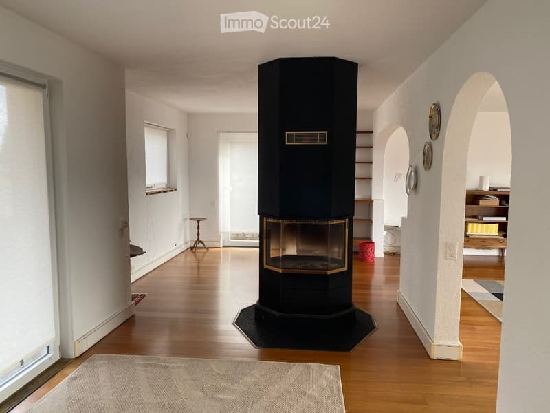 Spacious hallway with wooden flooring, white walls, and a central black fireplace with a gold trim. The hallway features arched doorways leading to adjacent rooms, with visible artwork on the walls and a small table near the window.