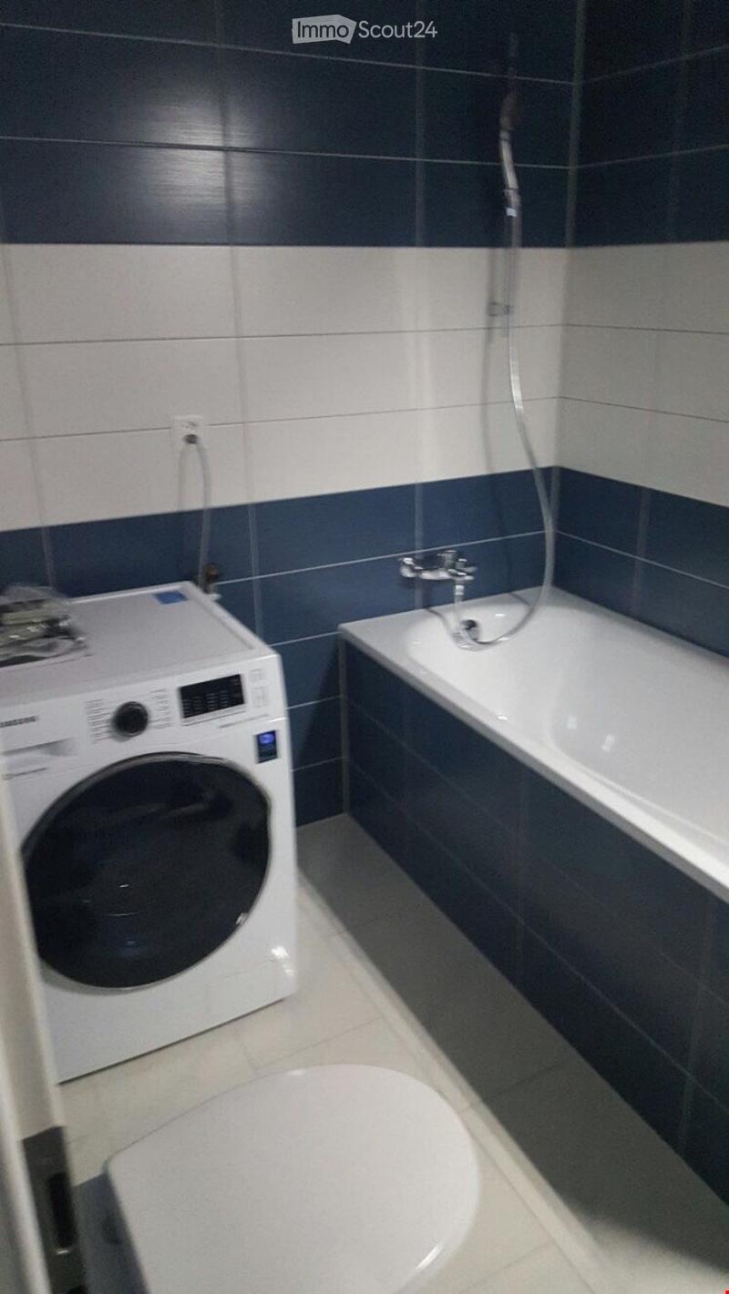 Modern bathroom with a bathtub, toilet, and a private washing machine. The walls are tiled in dark blue and white, and the floor is tiled in light gray.