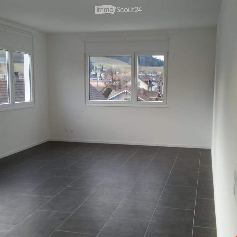 Empty room with grey tiled floor, white walls, and large windows offering a view of a residential area.