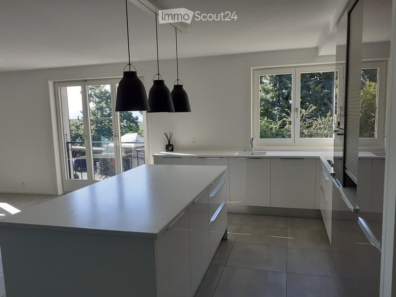 Modern kitchen with a large island, pendant lights, and white cabinets. Features a large window and a sliding glass door leading to a balcony.