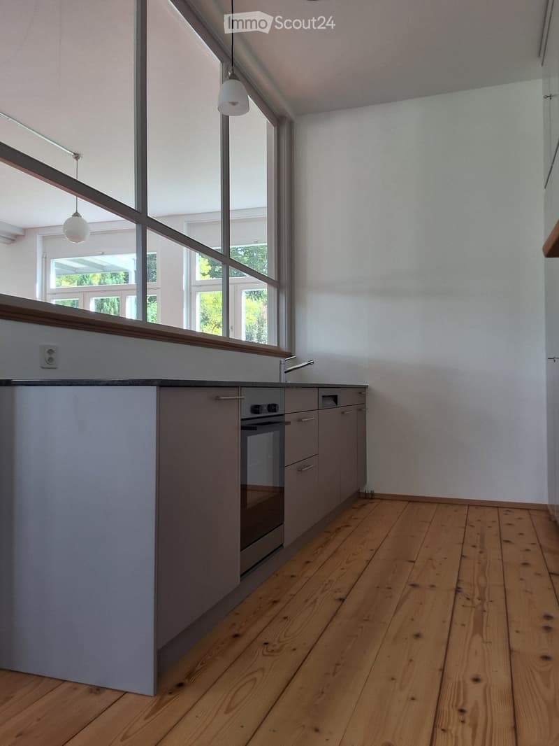 Modern kitchen with grey cabinets, wooden floor, large windows, and built-in oven.