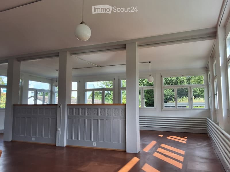 Empty room with wooden flooring, large windows, and decorative paneling on the lower walls. Natural light streams in, creating shadows on the floor.