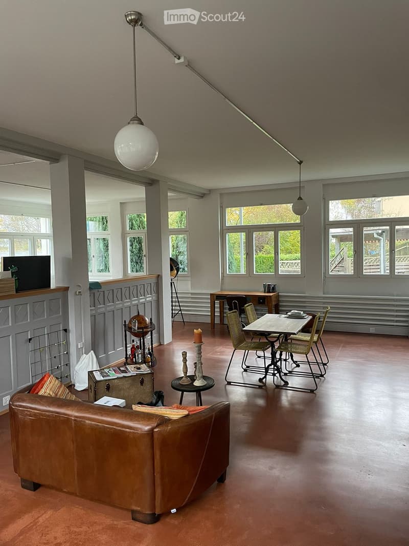 Spacious living room with wooden flooring, large windows, a brown leather sofa, a small table with chairs, a globe, and a TV in the background.