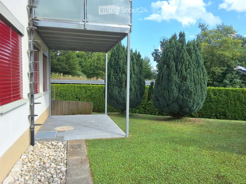 Well-maintained garden with a paved patio, neatly trimmed hedges, and tall coniferous trees. The patio features a covered walkway leading to the house, with a gravel border and a small grassy area.