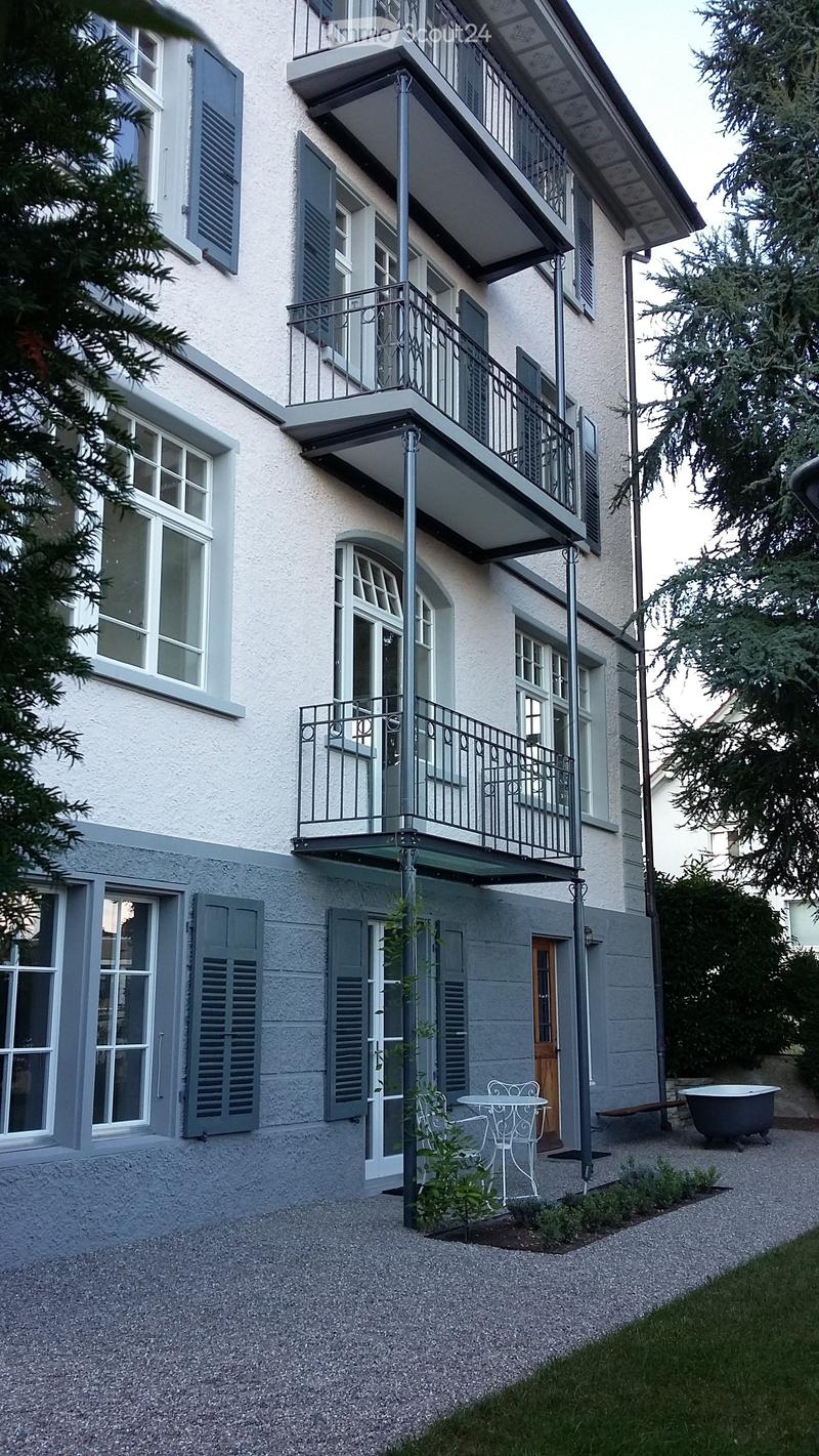 3 story building, balconies, white windows, grey shutters, table and chairs outside, plant