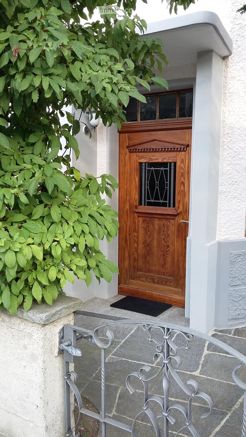 Entrance to a house with a wooden door, glass panels, and a decorative metal gate. The door is flanked by white walls and a small step leads up to it. Greenery is visible on the left side.
