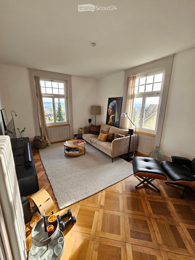 living room, hardwood floor, large windows, large beige couch with many pillows, coffee table with books, floor lamp, painting on wall, ottoman, black leather armchair