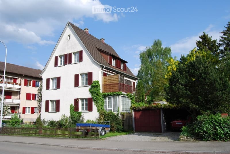 Two-story house with white walls, red shutters, and a balcony on the upper floor. Features a garage and a small trailer in the front yard.