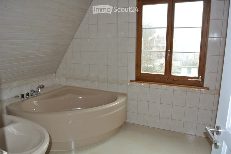 Bathroom with a large, curved bathtub, a sink, and a window with wooden frame. The walls are tiled, and the floor is light-colored.