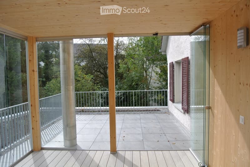 Modern balcony with wooden decking, glass railings, and a view of greenery outside.