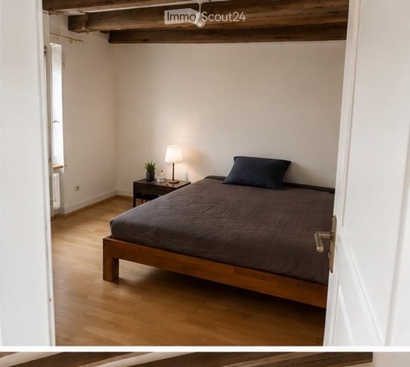 Cozy bedroom with wooden flooring, a single bed with dark bedding, a nightstand with a lamp and small plant, exposed wooden ceiling beams, and a partially open door leading to another room.