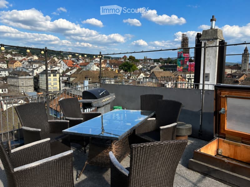 Roof terrace with outdoor furniture, including a table and chairs, overlooking a cityscape with buildings and a church tower.