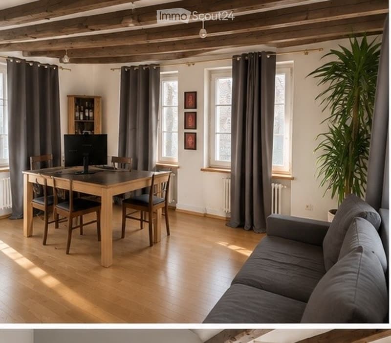 Bright, open-plan living and dining area with wooden flooring, large windows, grey sofa, wooden dining table with chairs, TV, cabinet, and a potted plant.