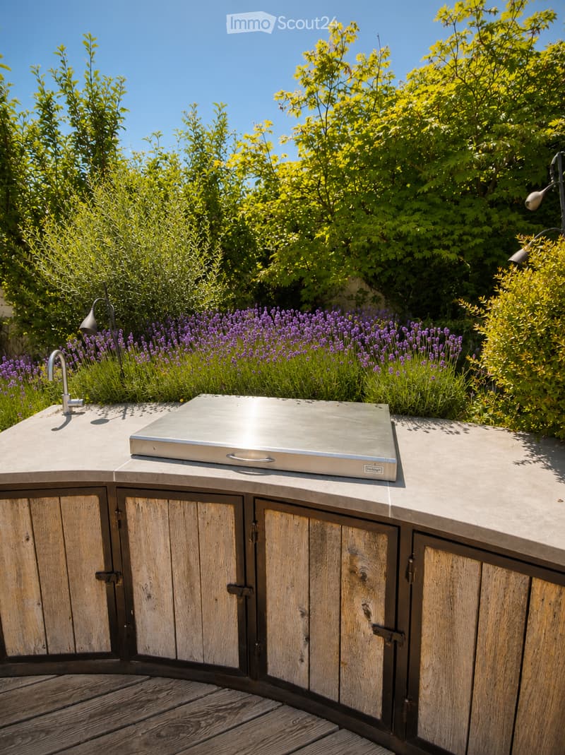 Outdoor kitchen area with wooden cabinetry, stainless steel grill, and surrounding greenery including lavender plants and trees.