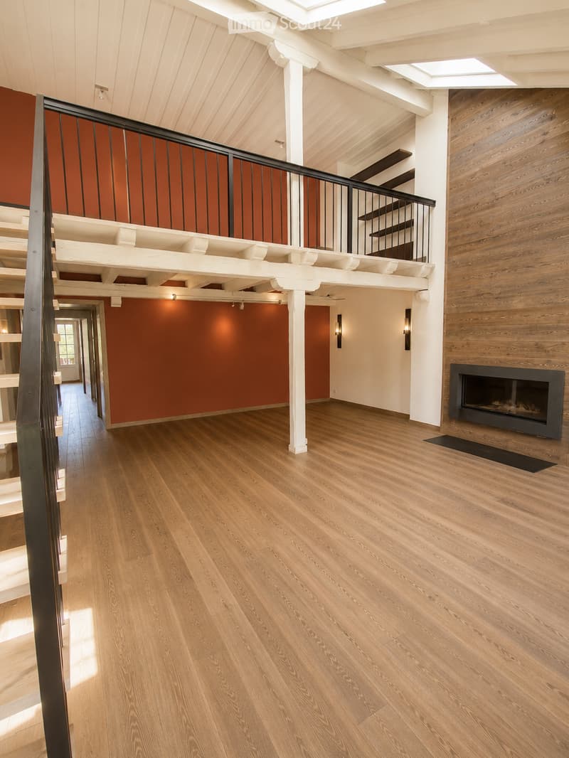 Spacious hallway with polished hardwood floors, white columns, and a black metal railing leading to an upper level. Features a built-in fireplace with a dark surround and warm lighting fixtures on the walls.