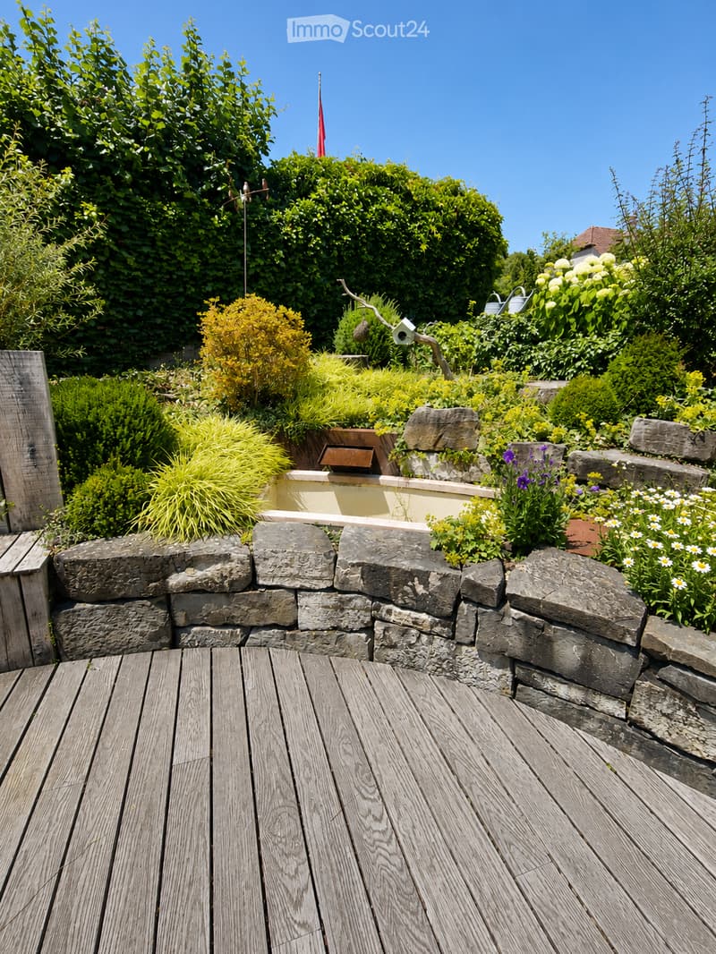 A wooden deck surrounded by a stone wall, with various plants, flowers, and shrubs in the background. The area is well-maintained and features a small birdhouse on a wooden post.