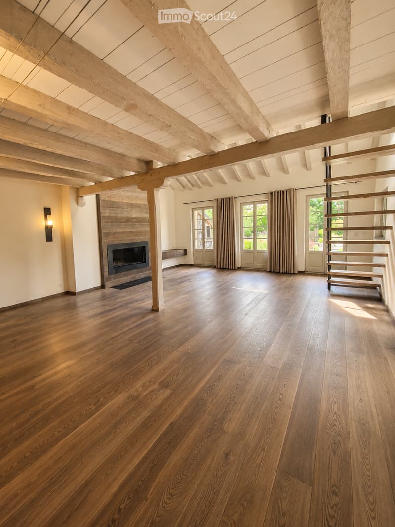 Open, spacious room with exposed wooden beams and polished hardwood flooring. Features a fireplace with a wooden surround, large windows with curtains, and a modern staircase leading upwards.