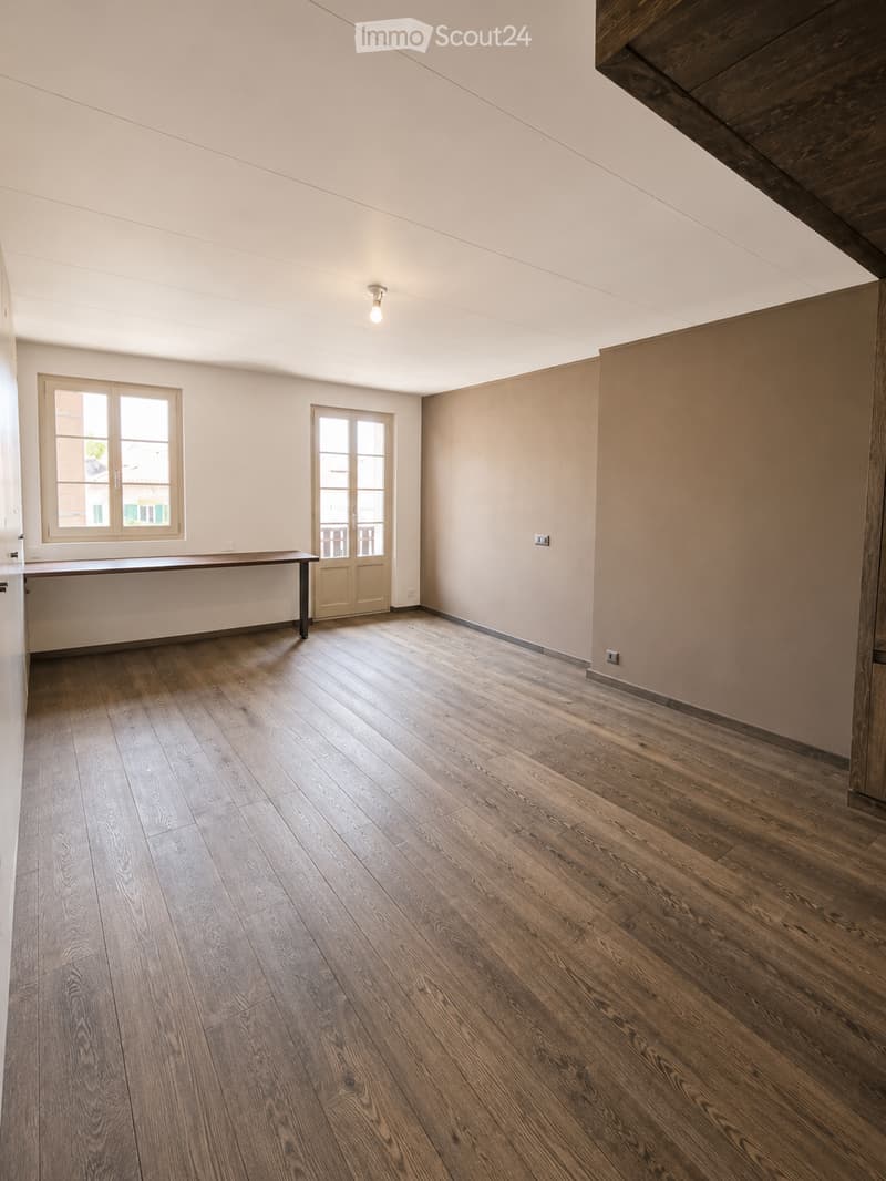 Empty room with wooden flooring, white walls, and large windows allowing natural light. A single table is positioned against one wall, and a door leads to another space.