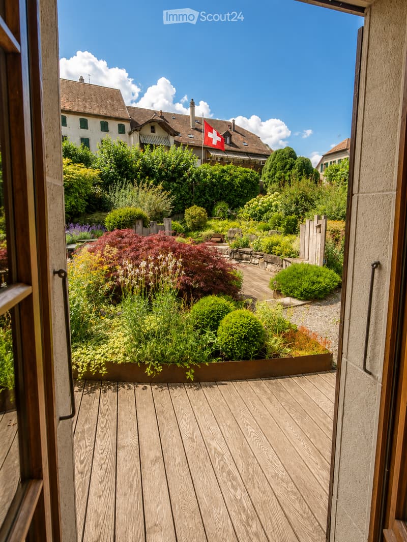 Wooden deck terrace with well-maintained garden, shrubs, and a view of a house with a Swiss flag.