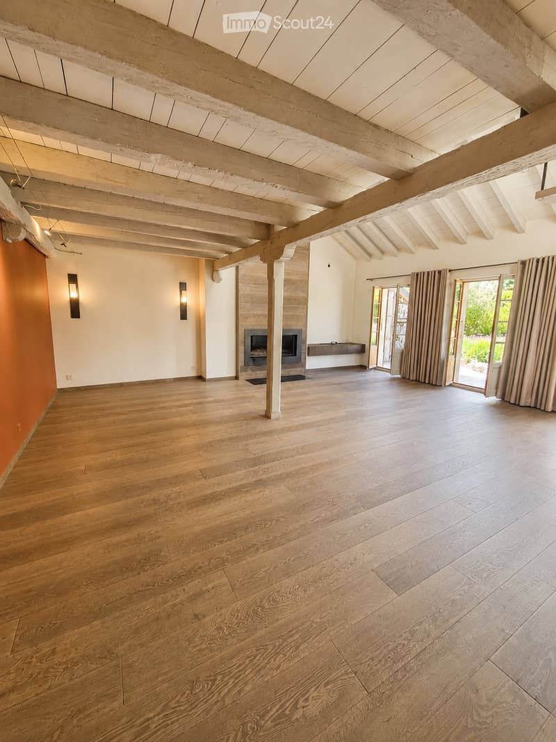 Spacious, unfurnished room with exposed wooden beams, hardwood flooring, and a fireplace. Large windows and sliding glass doors lead to an outdoor area.